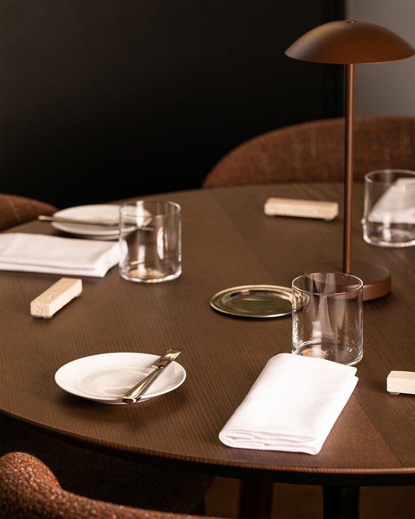 Il tavolo apparecchiato: piatti, bicchieri, tovaglioli piegati, segnaposto in legno e la lampada da tavolo color rame, Pulejo, Lion Brand