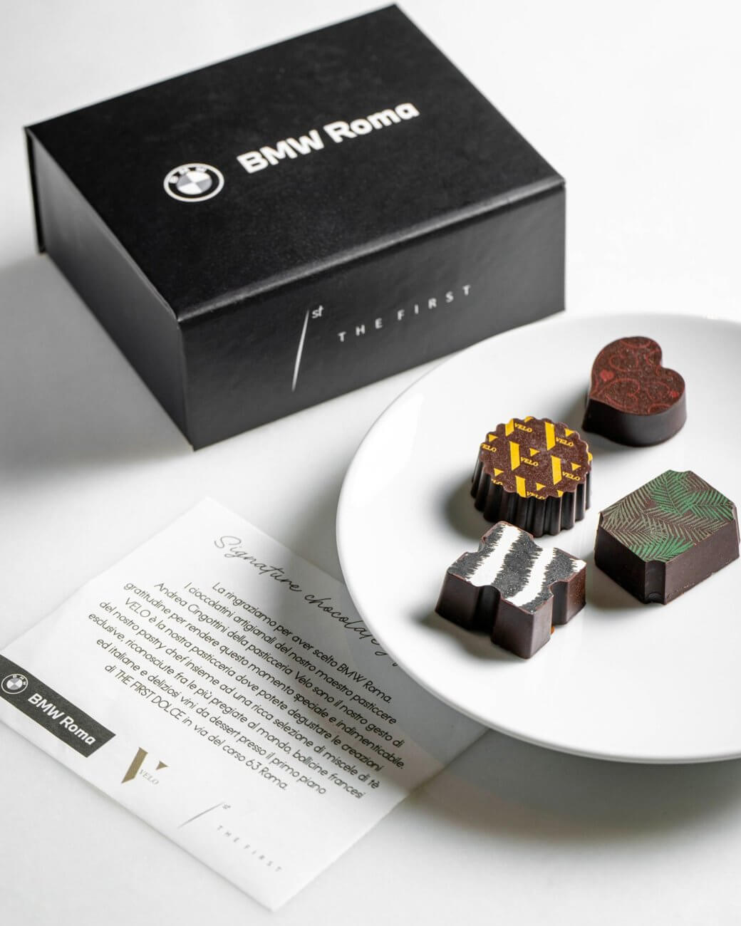 La scatola nera «BMW Roma / The First», un piatto con quattro cioccolatini decorati e la card «Signature tasting dinner», The First e BMW Roma, Lion Brand