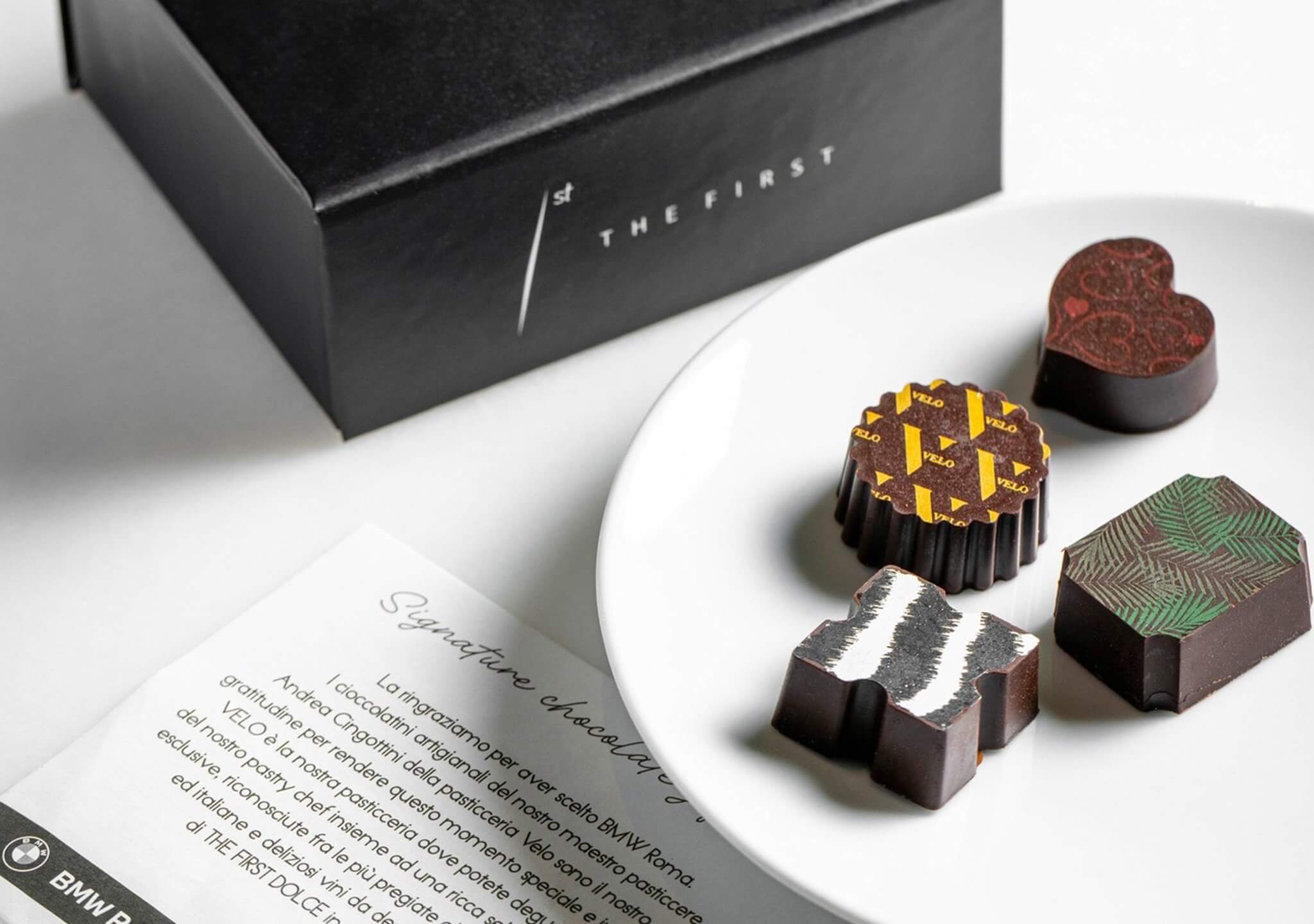 La scatola nera «BMW Roma / The First», un piatto con quattro cioccolatini decorati e la card «Signature tasting dinner», The First e BMW Roma, Lion Brand