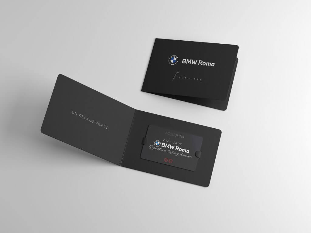 Il porta card nero aperto con la gift card inserita e, dietro, il porta card chiuso col logo, The First e BMW Roma, Lion Brand