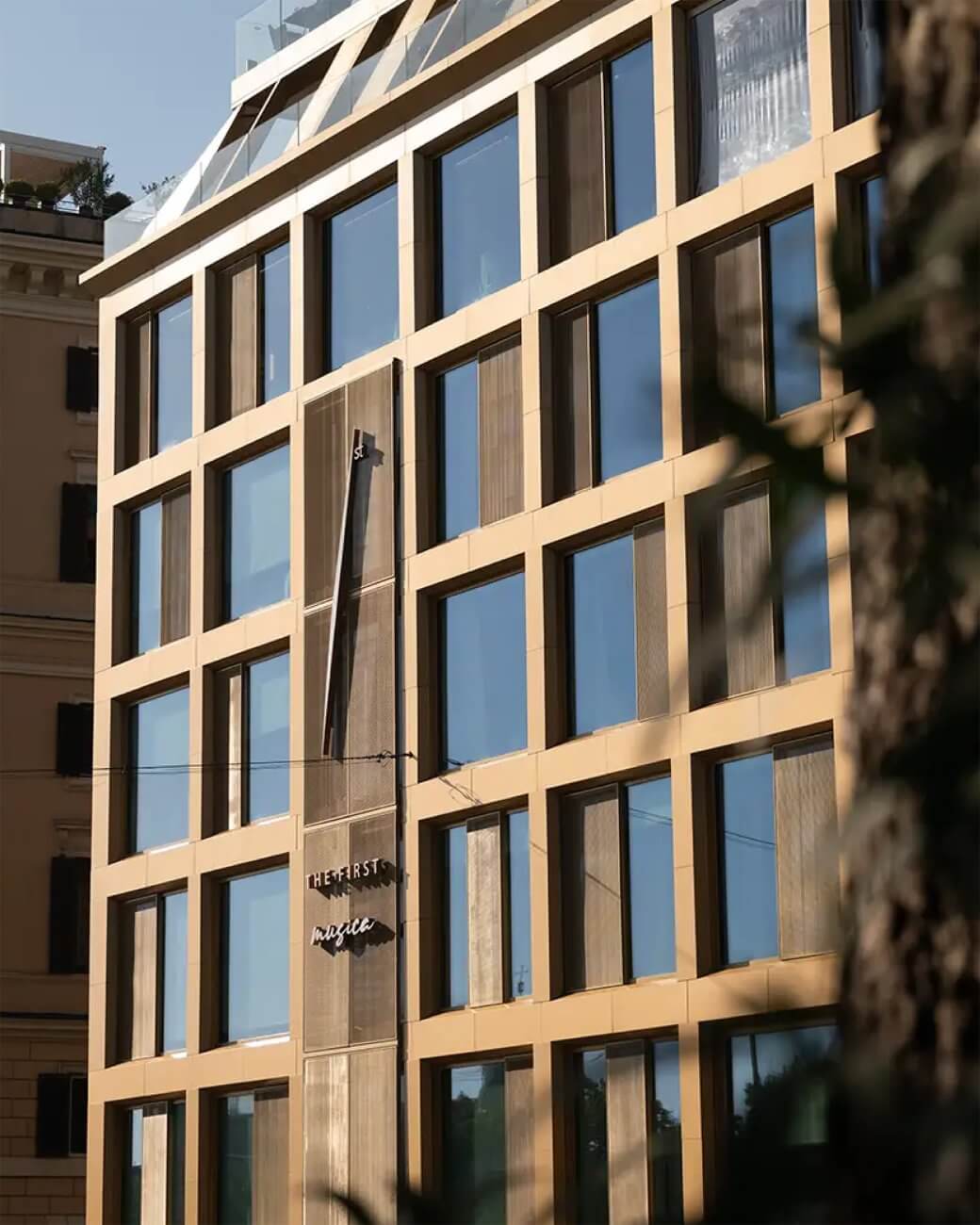 La facciata dell'hotel: la griglia di finestre e lesene color sabbia, l'insegna sulla porta, un ramo in primo piano, The First Musica, Lion Brand