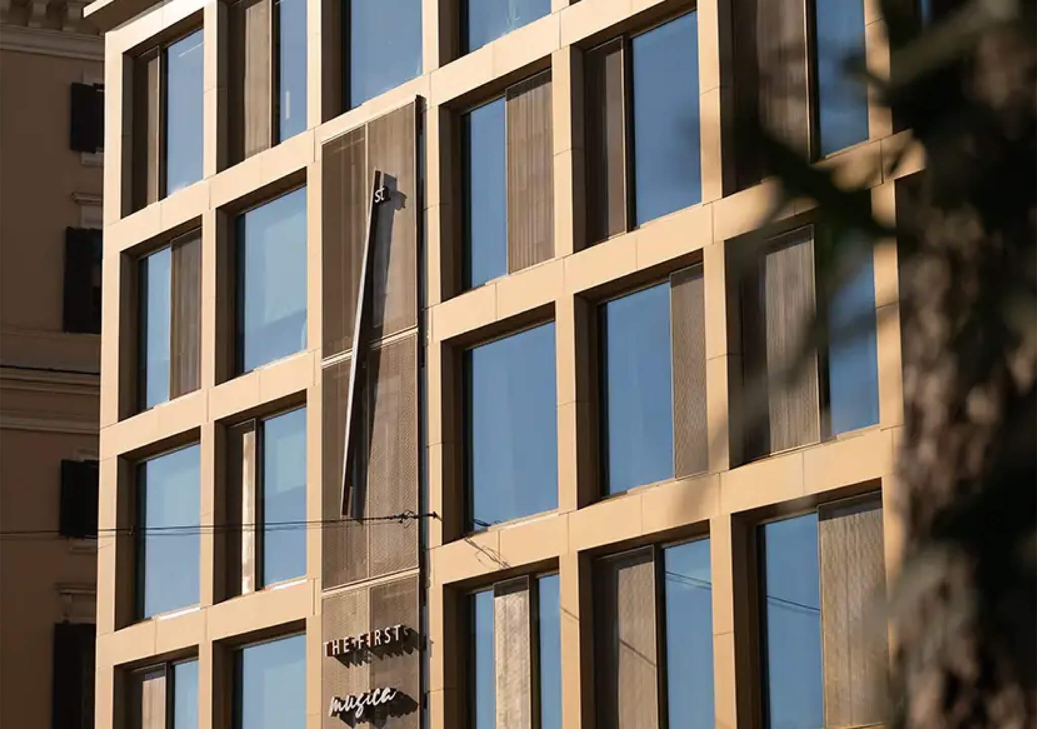 La facciata dell'hotel: la griglia di finestre e lesene color sabbia, l'insegna sulla porta, un ramo in primo piano, The First Musica, Lion Brand
